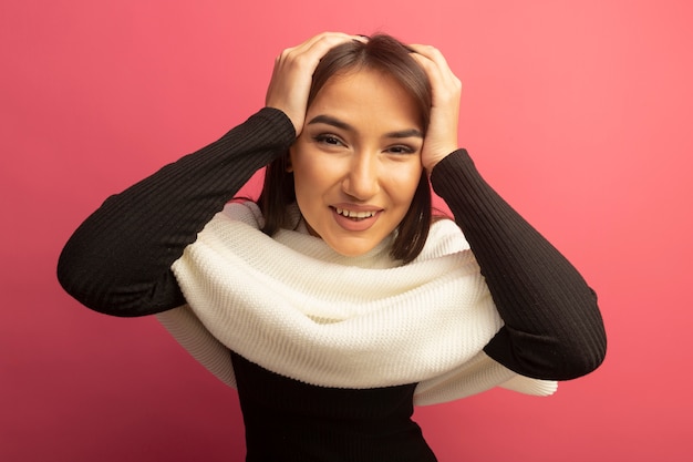 Foto gratuita mujer joven con pañuelo blanco mirando al frente con la mano en la cabeza sonriendo alegremente de pie sobre la pared rosa