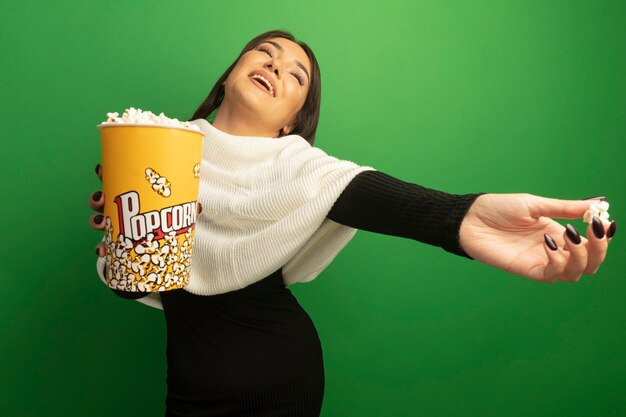 Mujer joven con pañuelo blanco mostrando cubo con palomitas de maíz feliz y positivo sonriendo alegremente con el brazo extendido