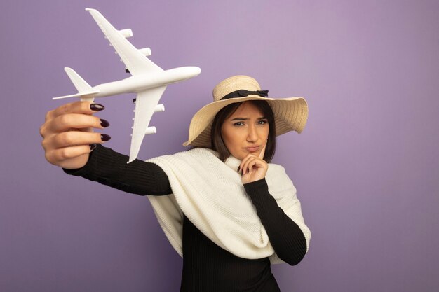 Mujer joven con pañuelo blanco en sombrero de verano mostrando avión de juguete con la mano en la barbilla pensando con cara seria