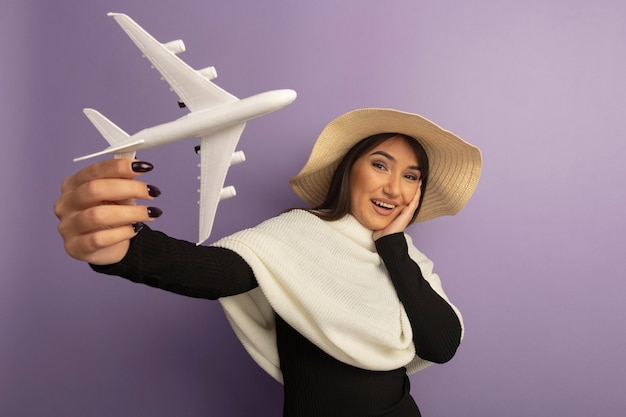 Mujer joven con pañuelo blanco en sombrero de verano mostrando avión de juguete sonriendo feliz y alegre