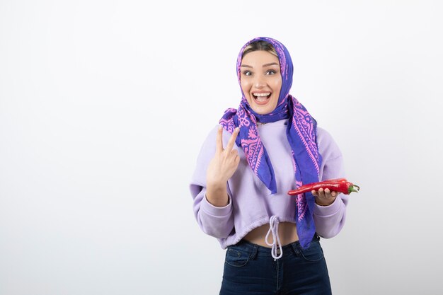 Foto gratuita mujer joven en pañuelo sosteniendo un pimiento rojo y mostrando el signo de la victoria.