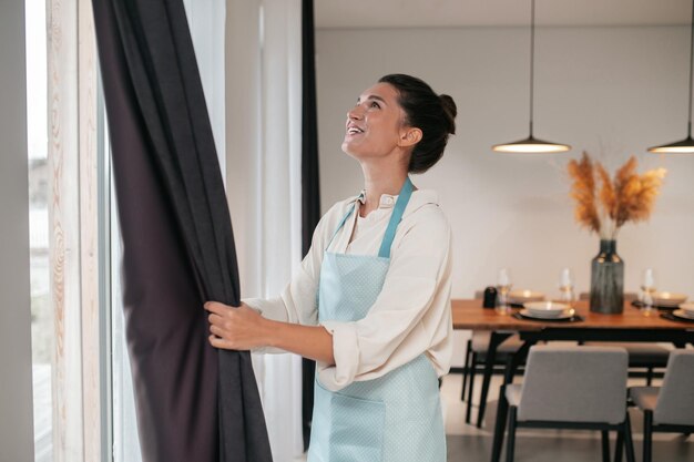 Mujer joven parada cerca de la viuda y arreglando las cortinas