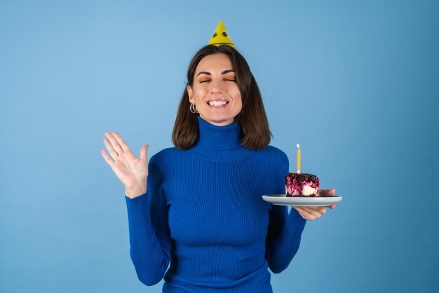 Mujer joven en una pared azul celebra un cumpleaños, sostiene un trozo de tarta, de muy buen humor