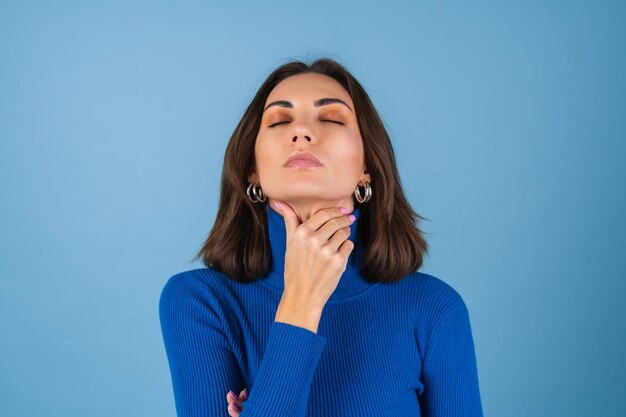 Mujer joven en una pared azul toca su garganta, sufre de dolor
