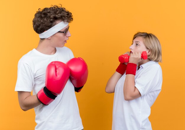 Mujer joven pareja deportiva con pesas mirando a su novio con guantes de boxeo sorprendido y confundido de pie sobre la pared naranja