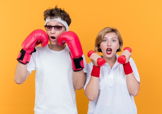 Mujer joven pareja deportiva con pesas de pie junto a su novio con guantes de boxeo posando sorprendido y confundido de pie sobre la pared naranja
