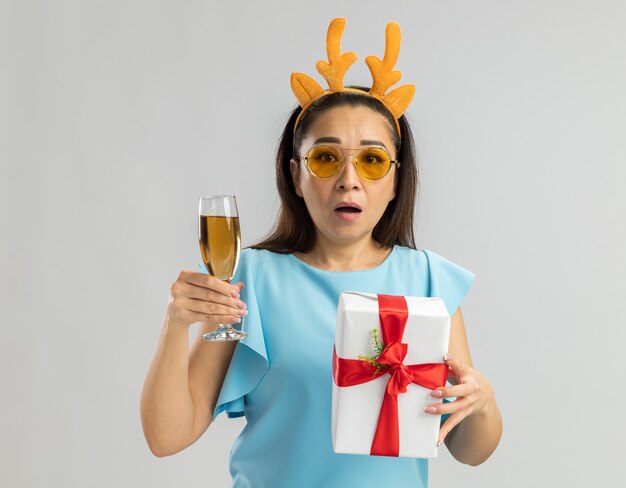 Mujer joven en la parte superior azul con borde divertido con cuernos de venado y gafas amarillas sosteniendo una copa de champán y regalo de Navidad con aspecto preocupado y confundido