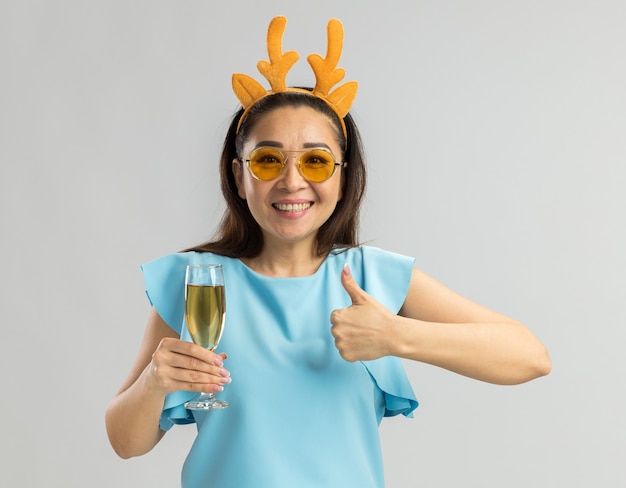 Mujer joven en la parte superior azul con borde divertido con cuernos de venado y vasos amarillos sosteniendo una copa de champán mirando feliz y alegre sonriendo mostrando los pulgares para arriba
