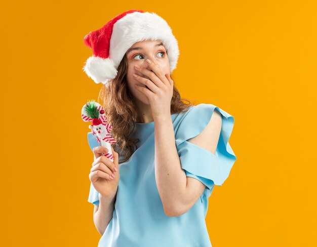 Mujer joven en la parte superior azul y gorro de Papá Noel con bastón de caramelo de Navidad mirando a un lado conmocionado cubriendo la boca con la mano