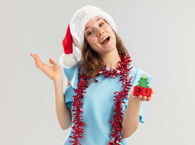 Mujer joven en la parte superior azul y gorro de Papá Noel con oropel alrededor de su cuello sosteniendo cubos de juguete con fecha de año feliz ney mirando feliz y positivo sonriendo