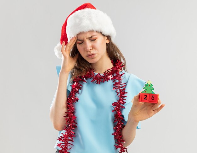 Mujer joven en la parte superior azul y gorro de Papá Noel con oropel alrededor de su cuello sosteniendo cubos de juguete con fecha de Navidad mirando desagradable con fuerte dolor de cabeza tocando su cabeza