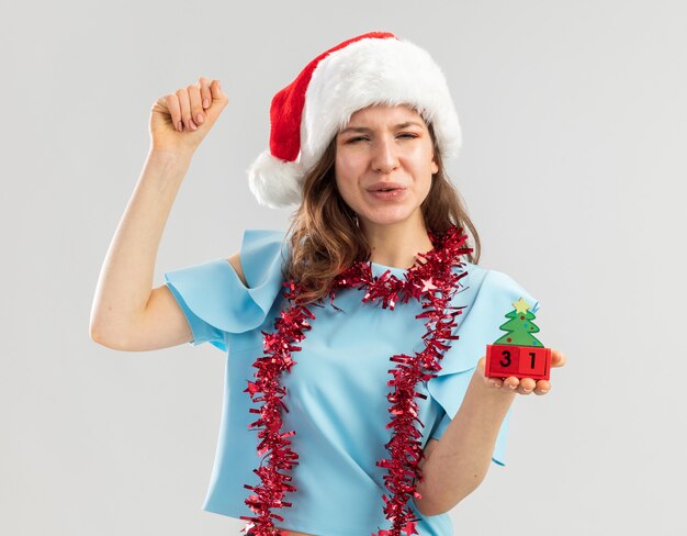 Mujer joven en la parte superior azul y gorro de Papá Noel con oropel alrededor de su cuello sosteniendo cubos de juguete con feliz año ney fecha apretando el puño feliz y emocionado