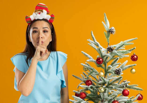 Mujer joven en la parte superior azul con gracioso borde navideño en la cabeza haciendo gesto de silencio con el dedo en los labios de pie junto a un árbol de Navidad sobre la pared naranja