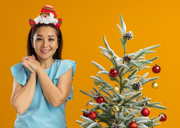 Mujer joven en la parte superior azul con gracioso borde navideño en la cabeza mirando a cámara feliz y positivo tomados de la mano juntos sonriendo alegremente de pie junto a un árbol de Navidad sobre fondo naranja