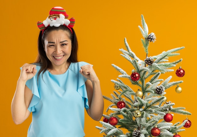 Mujer joven en la parte superior azul vistiendo gracioso borde navideño en la cabeza apretando los puños feliz y emocionado de pie junto a un árbol de Navidad sobre fondo naranja