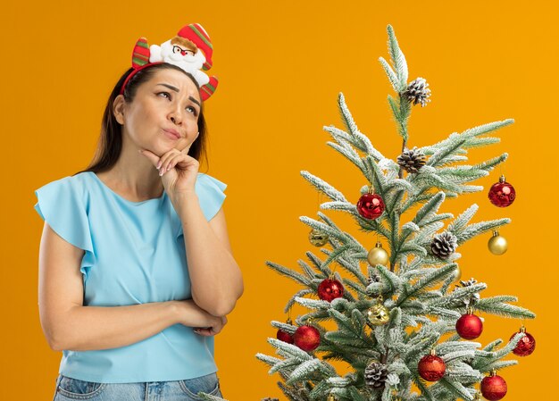 Mujer joven en la parte superior azul vistiendo gracioso borde navideño en la cabeza de pie junto a un árbol de navidad mirando hacia arriba desconcertado sobre fondo naranja