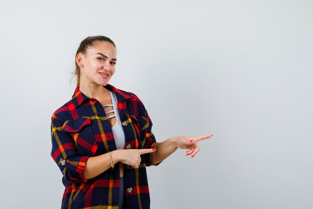 Mujer joven en la parte superior de la cosecha, camisa a cuadros apuntando hacia el lado derecho y mirando alegre, vista frontal.