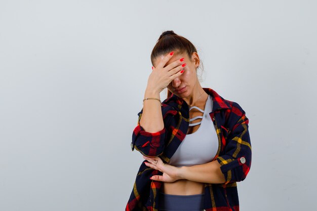 Mujer joven en la parte superior de la cosecha, camisa a cuadros, pantalones con la mano en la frente y mirando exhausto, vista frontal.