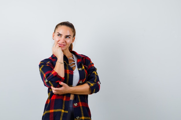 Mujer joven en la parte superior de la cosecha, camisa a cuadros de pie en pose de pensamiento y mirando pensativo, vista frontal.