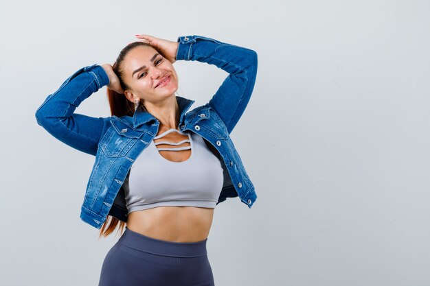 Mujer joven en la parte superior de la cosecha, chaqueta, pantalón con las manos en la cabeza y mirando alegre, vista frontal.