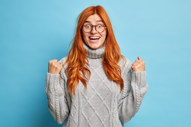 Mujer joven pelirroja alegre con expresión alegre se regocija el éxito aprieta los puños después de los objetivos de logro vestida con suéter gris de punto.