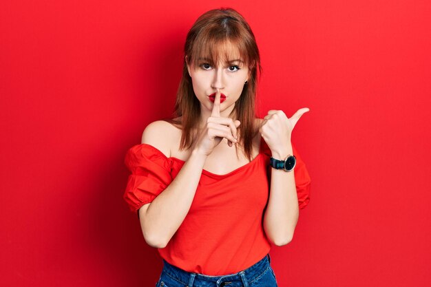 Mujer joven pelirroja con una camiseta roja informal que pide silencio con el dedo en los labios apuntando con la mano al lado del silencio y el concepto secreto