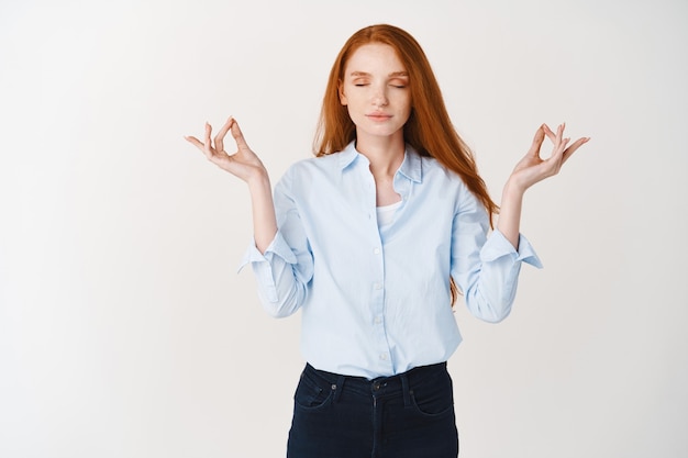 Mujer joven pelirroja que mantiene la calma con la meditación, cierra los ojos y extiende las manos en el signo mudra om, practica yoga contra la pared blanca