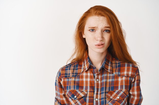 Mujer joven pelirroja que parece dudosa e incómoda, que tiene dudas, de pie sobre una pared blanca