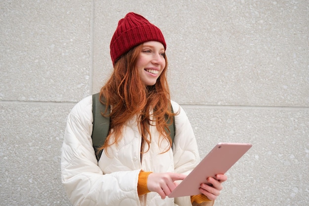 Mujer joven pelirroja con sombrero rojo usa su tableta digital al aire libre se encuentra en la calle con gadget cone