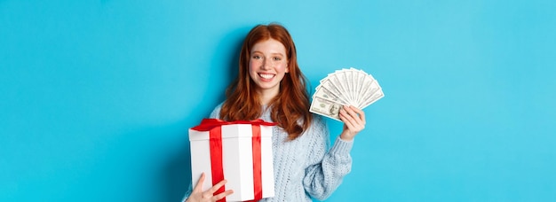 Foto gratuita mujer joven pelirroja sosteniendo caja de regalo de navidad y dinero sonriendo complacido de pie sobre backgrou azul
