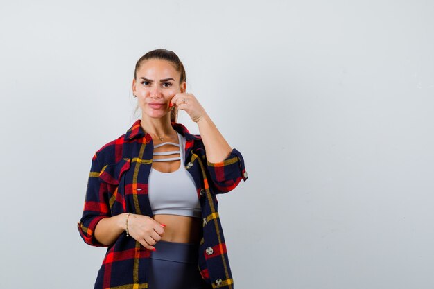 Mujer joven pellizcando la mejilla en la parte superior de la cosecha, camisa a cuadros y luciendo linda. vista frontal.