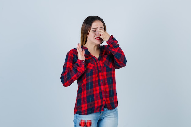 Mujer joven pellizcando la nariz, mostrando la señal de pare mientras llora en camisa a cuadros, jeans y luciendo malhumorado. vista frontal.