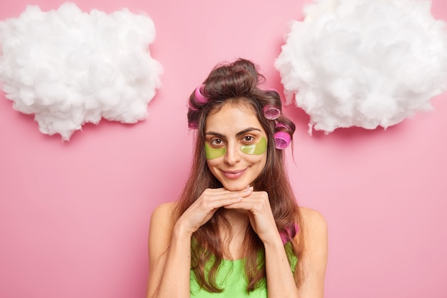 Mujer joven de pelo bastante oscuro con rizadores de pelo hace peinado aplica parches de colágeno debajo de los ojos para reducir las sonrisas de hinchazón agradablemente aisladas sobre la pared rosa