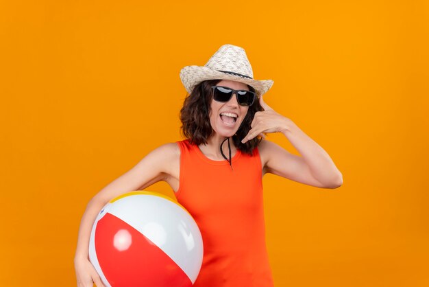 Una mujer joven con el pelo corto en una camisa naranja con sombrero para el sol y gafas de sol sosteniendo una bola inflable mostrando el gesto de llamarme