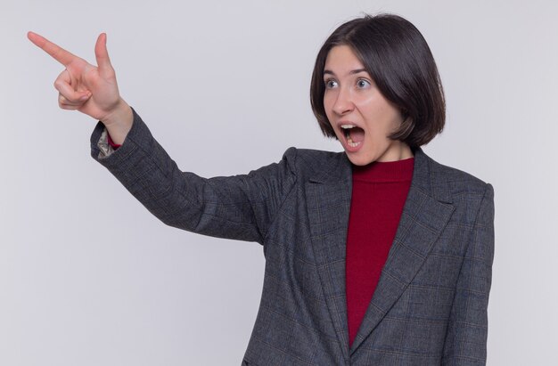 Mujer joven con pelo corto con chaqueta gris mirando a un lado sorprendido y confundido apuntando con el dedo índice a algo parado sobre la pared blanca