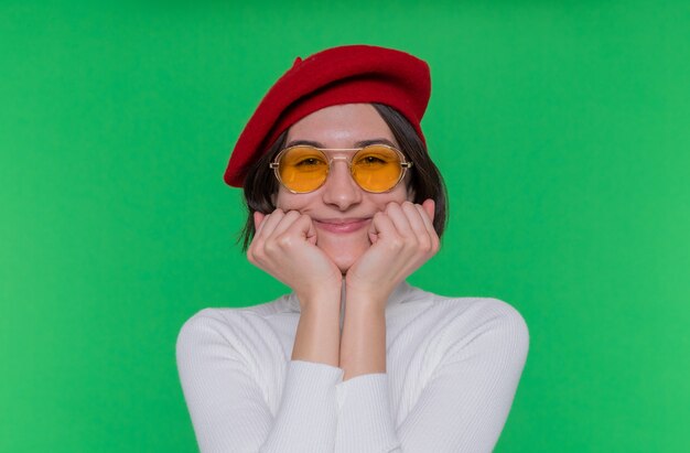 Mujer joven con pelo corto en cuello alto blanco con boina y gafas de sol amarillas mirando al frente feliz y positivo sonriendo alegremente de pie sobre la pared verde