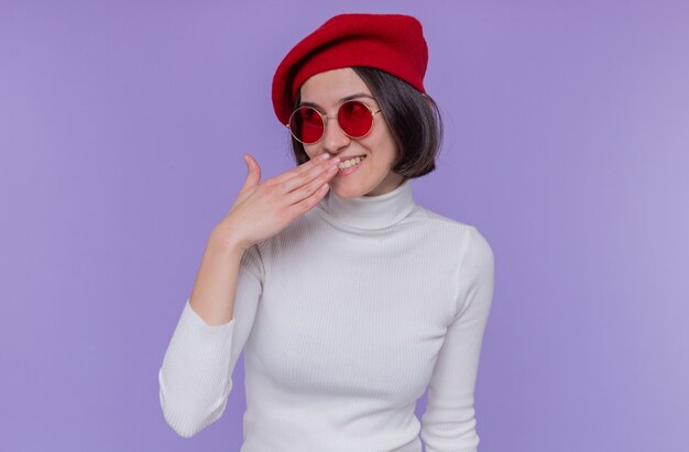 Mujer joven con pelo corto en cuello alto blanco con boina y gafas de sol rojas mirando feliz