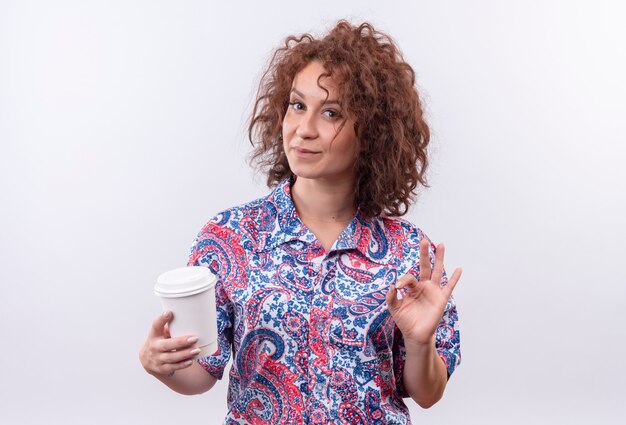 Mujer joven con pelo corto y rizado en camisa colorida sosteniendo la taza de café mostrando signo ok sonriendo de pie sobre la pared blanca