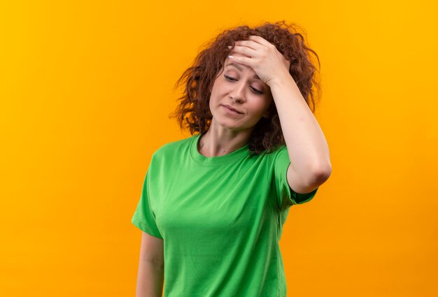 Mujer joven con pelo corto y rizado en camiseta verde con aspecto cansado y aburrido con la mano en la cabeza con dolor de cabeza de pie