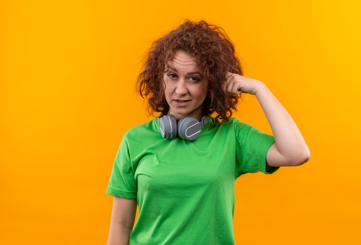 Foto gratuito mujer joven con pelo corto y rizado en camiseta verde con auriculares mirando confundido señalando con el dedo su sien sobre pared naranja