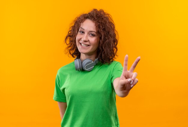 Mujer joven con pelo corto y rizado en camiseta verde con auriculares mostrando el signo de la victoria sonriendo alegremente de pie sobre la pared naranja