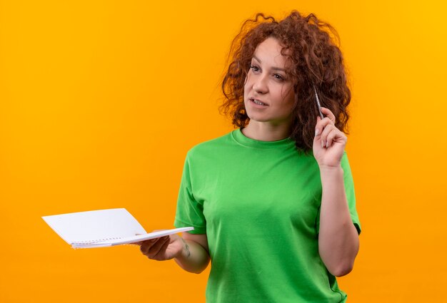 Mujer joven con pelo corto y rizado en camiseta verde con cuaderno y bolígrafo mirando a un lado con expresión pensativa en la cara de pie