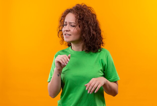 Mujer joven con pelo corto y rizado en camiseta verde haciendo gesto de defensa con expresión de disgusto de pie sobre la pared naranja