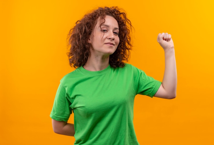 Foto gratuito mujer joven con pelo corto y rizado en camiseta verde levantando el puño mirando confiado de pie sobre la pared naranja