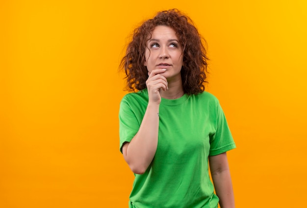 Mujer joven con pelo corto y rizado en camiseta verde mirando hacia arriba con expresión pensativa en la cara pensando de pie sobre la pared naranja