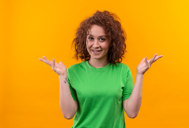 Foto gratuito mujer joven con pelo corto y rizado en camiseta verde mirando positivo y feliz extendiendo los brazos a los lados de pie