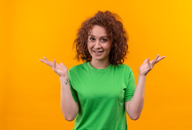 Mujer joven con pelo corto y rizado en camiseta verde mirando positivo y feliz extendiendo los brazos a los lados de pie