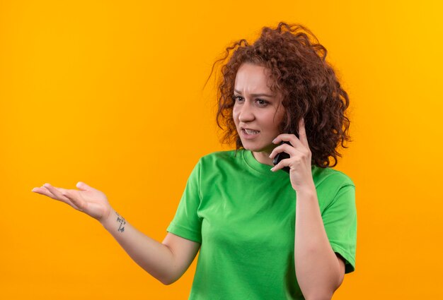 Mujer joven con pelo corto y rizado en camiseta verde que parece confundida y muy ansiosa mientras habla por teléfono móvil de pie