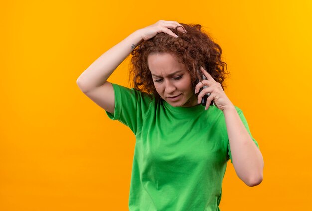 Mujer joven con pelo corto y rizado en camiseta verde que parece confundida y muy ansiosa mientras habla por teléfono móvil de pie