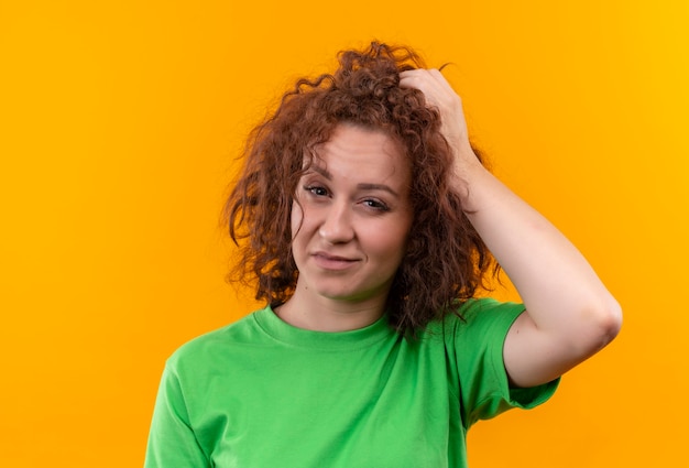 Mujer joven con pelo corto y rizado en camiseta verde que parece confundida y muy ansiosa tocando su cabeza de pie